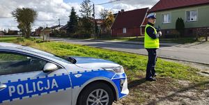 Radiowóz, dalej  przy drodze stoi policjant, który miernikiem mierzy prędkość.