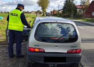 Policjant trzyma w ręku urządzenie akoblow, stoi przy zatrzymanym do kontroli pojeździe.