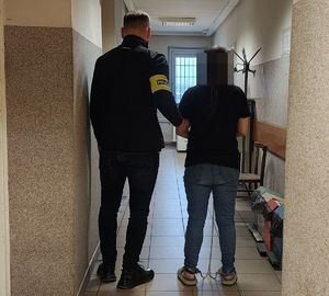 Policjant prowadzi zatrzymaną po korytarzu w budynku komendy.