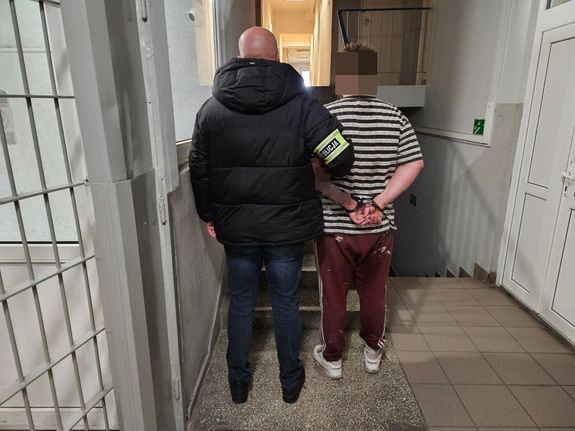 Policjant prowadzi zatrzymanego po korytarzu w budynku komendy.