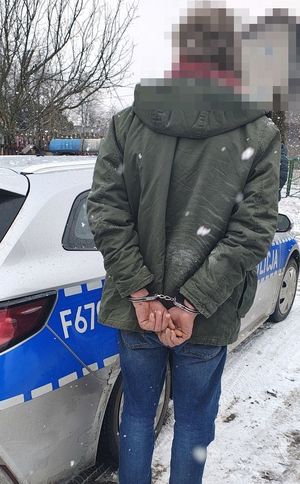 Osoba zatrzymana stoi obok radiowozu ma kajdanki założone na rękach.