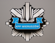 Odznaka policyjna w kolorze szarym. W środku na niebieskiej szarfie napis &quot;KPP Wieruszów&quot;.