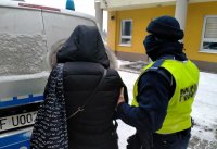 Umundurowany policjant prowadzi zatrzymaną. W tle oznakowany radiowóz oraz budynek KPP w Wieruszowie.