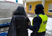 Umundurowany policjant prowadzi zatrzymaną. W tle oznakowany radiowóz oraz budynek KPP w Wieruszowie.
