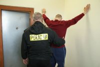 Umundurowany policjant przeszukuje zatrzymanego w pomieszczeniu dla osób zatrzymanych.