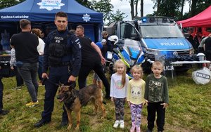 widzimy dzieci stojących z policjantem oraz psem służbowym na tle policyjnego radiowozu.