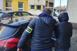 widzimy policjanta z zatrzymana kobieta na tle pojazdu i budynku komendy.