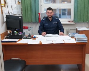 widzimy policjanta siedzącego za biurkiem trzymający w ręku długopis.