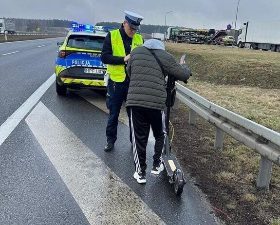 widzimy policjanta kontrolującego mężczyznę-kierującego hulajnogą elektryczną, w tle widać radiowóz.