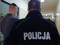 policjant prowadzi zatrzymanego. mężczyźni stoją tyłem. Zdjęcie uwidacznia ich postacie od głowy do pasa. Policjant umundurowany , na plecach napis POLICJA. Zatrzymany ubrany w bluzę koloru szarego, widać, że ma założoną maseczkę zasłaniającą usta i nos. Zdjęcie wykonane w pomieszczeniu służbowym komendy policji.