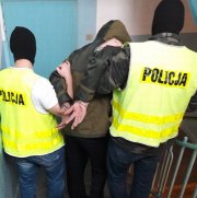 policjanci z wydziału kryminalnego ubrani po cywilnemu w policyjnych kamizelkach z kominiarkami na głowie, prowadzą zatrzymanego mężczyzne