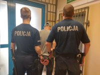 policjanci umundurowani prowadzą zatrzymanego, mężczyzna ma kajdanki założone na dłonie trzymane z tyłu