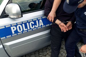 na zdjęciu widoczny radiowóz policyjny oraz częściowo zatrzymany mężczyzna który ma kajdanki założone na ręce trzymane z tyłu.