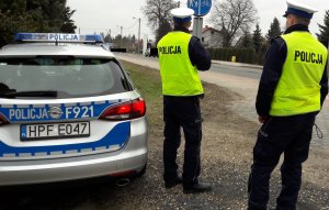 policjanci umundurowani stoją przy drodze obok radiowozu. w tle ulica i obszar zabudowany