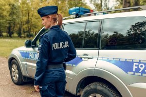 policjantka stoi przy radiowozie policyjnym. w tle zieleń, drzewa