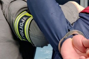 na zdjęciu fragment dłoni policjanta i osoby zatrzymanej, która ma założone kajdanki na ręce trzymane z tyłu