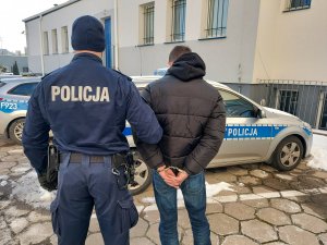 umundurowany policjant prowadzi zatrzymanego, który ma założone kajdanki na ręce trzymane z tyłu. w tle radiowozy policyjne które stoją na placu wewnętrznym komendy policji