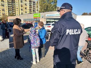 akcja odblaskowa pod nazwa świeć przykładem. Na zdjęciu komendant powiatowy policji w Bełchatowie inspektor Grzegorz Czubakowski  którego widać z tyłu. Na zdjęciu  Starosta Bełchatowski Dorota Pędziwiatr która dwójce dzieciom wręcza elementy odblaskowe. w tle osiedlowe bloki i sklepy oraz zaparkowane na parkingu auta.