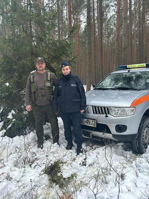 Policjantka wraz ze strażnikiem Straży Leśnej patrolują kompleks leśny. Obok nich stoi pojazd Straży Leśnej