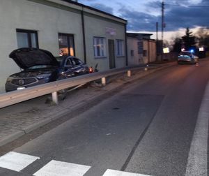 Po lewej stronie na chodniku stoi uszkodzony pojazd w oddali zaparkowany radiowóz