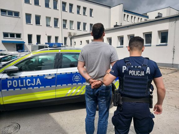 umundurowany policjant prowadzi zatrzymanego mężczyznę. zdjęcie zdobione na placu wewnętrznym komendy policji. w tle radiowozy i budynek komendy