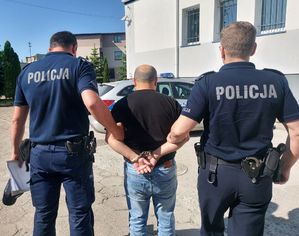 umundurowani policjanci prowadzą zatrzymanego mężczyznę. zdjęcie zdobione na placu wewnętrznym komendy policji. w tle radiowozy