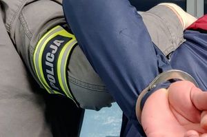 Na zdjęciu ręka policjanta nieumundurowanego z opaską policja oraz dłonie mężczyzny, który na ręce trzymane z tyłu ma założone kajdanki.