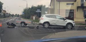 miejsce zdarzenia, skrzyżowanie ulic, samochód osobowy z podpiętą przyczepką, na drodze leży motocykl, w tle ulice i domy.