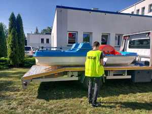 policjant w czasie oględzin odzyskanego przez policjantów skradzionego roweru wodnego. Sprzęt stoi na pojeździe typu laweta. Zdjęcie zrobione na placu wewnętrznym komendy policji. W tle widoczny budynek i pojazdy służbowe.