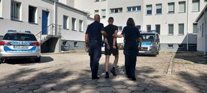 policjanci prowadzą zatrzymanego mężczyznę. zdjęcie zrobione na placu wewnętrznym komendy, w tle budynek policji oraz radiowozy.