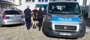 policjanci prowadzą zatrzymanego mężczyznę. zdjęcie zrobione na placu wewnętrznym komendy, w tle budynek policji oraz radiowozy.