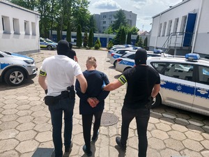 dwóch policjantów ubranych po cywilnemu prowadzi zatrzymanego mężczyznę. zdjęcie zrobione na placu wewnętrznym komendy policji, w tle radiowozy policyjne.