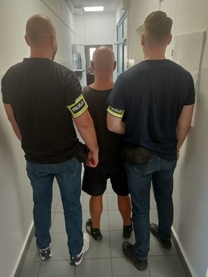 dwóch nieumundurowanych policjantów z założoną na przedramieniu opaską policja prowadzi zatrzymanego mężczyznę. Zdjęcie zrobione na korytarzu komendy policji.
