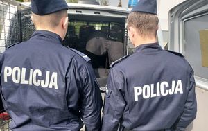dwóch umundurowanych policjantów, dalej widoczny tył radiowozu, w miejscu dla osób zatrzymanych widoczna osoba.