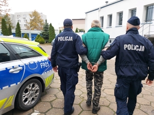 dwóch umundurowanych policjantów prowadzi zatrzymanego mężczyznę, zdjęcie zrobione na placu wewnętrznym komendy, w tle radiowozy policyjne i budynki.