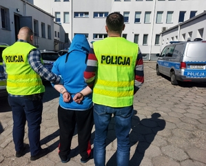 policjanci po cywilnemu z założonymi kamizelkami z napisem na plecach POLICJA prowadzą zatrzymanego mężczyznę, który na ręce trzymane z tyłu ma założone kajdanki. zdjęcie zrobione na placy wewnętrznym komendy, stoją tutaj radiowozy w tle widczny budynek komendy policji.