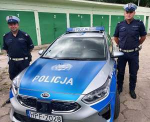 dwaj umundurowani policjanci referatu ruchu drogowego stoją przy oznakowanym radiowozie