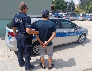 umundurowany policjant prowadzi zatrzymanego mężczyznę. W tle oznakowane radiowozy policyjne.