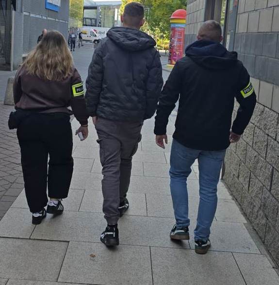 nieumundurowani policjanci z opaskami z napisem Policja na rękach prowadzą zaginionego mężczyznę.