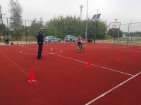 Zdjęcie przedstawia dziecko jadące na rowerze po torze, pod nadzorem policjanta. W tle widać osobę czekającą na przystąpienie egzaminu, boisko sportowe ogrodzone wysoką siatką, pachołki, tyczki  treningowe, kosz do koszykówki, drzewa, krzewy, dwa radiowozy.