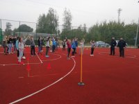 Zdjęcie przedstawia dwóch policjantów na boisku sportowym z grupą dzieci. Dzieci trzymają w rękach elementy odblaskowe. W tle zdjęcia widać boisko sportowe ogrodzone wysoką siatką, pachołki, tyczki  treningowe, drzewa, znaki drogowe, krzewy, dwa autobusy, trzy rowery, dwa radiowozy.