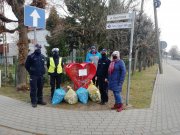 Na zdjęciu widać trzech umundurowanych policjantów oraz kobietę i mężczyznę, którzy stoją przy czerwonym pojemniku w kształcie serca, obok stoją cztery worki foliowe z plastikowymi nakrętkami. W tle widać budynek, ogrodzenie, znaki drogowe, drzewa i krzewy.