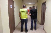 Korytarz komendy policji w Zduńskiej Woli. Policjant  ubrany w żółtą odblaskową kamizelkę prowadzi zatrzymanego.