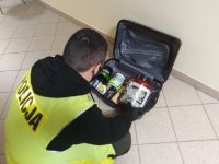 Policjant ubrany w żółtą kamizelkę odblaskową otwiera walizkę, w której są różne pojemniki z narkotykami