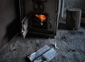 Na zdjęciu piec w którym pali się ogień, obok leży gazeta i kłoda drewna