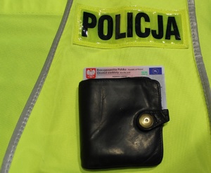 Czarny portfel i dowód osobisty, leżą na kamizelce odblaskowej z napisem &quot;Policja&quot;.