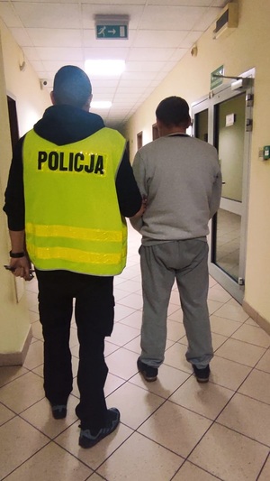 Policjant ubrany w kamizelkę odblaskową z napisem &quot;POLICJA&quot; prowadzi osobę zatrzymaną.
