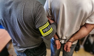 Policjant trzyma za ramię zatrzymanego, który ma kajdanki na ręce trzymane z tyłu.