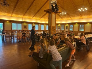 Sala, stoją w rzędach stoły i krzesła, dzieci siedzą na sofach i krzesłach, zebrały się wokół policjantki, która mówi im o bezpieczeństwie