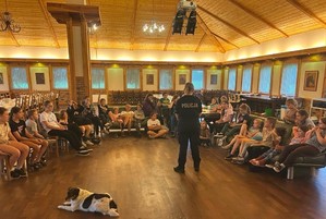 Sala, stoją w rzędach stoły i krzesła, dzieci siedzą na sofach i krzesłach, zebrały się wokół policjantki, która mówi im o bezpieczeństwie